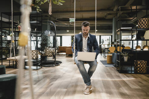 Geschäftsmann mit Laptop im Coworking Space, auf einer Schaukel sitzend - KNSF06996