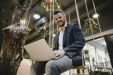 Geschäftsmann mit Laptop im Coworking Space, auf einer Schaukel sitzend - KNSF06998