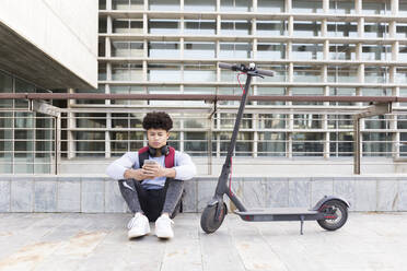 Young man with e-scooter relaxing and using smartphone - JPTF00390