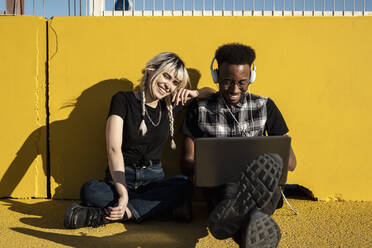 Porträt eines lachenden jungen Paares, das mit Kopfhörern und Laptop vor einer gelben Wand sitzt - RCPF00162