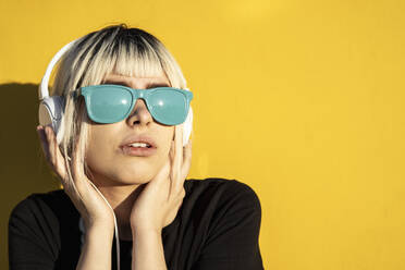 Porträt einer jungen Frau mit blauer Sonnenbrille, die vor einer gelben Wand Musik hört - RCPF00186