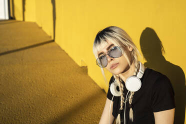 Porträt einer jungen Frau mit Kopfhörern und farbiger Sonnenbrille vor einer gelben Wand - RCPF00187