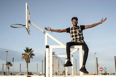 Portrait of young man sitting on soccer goal against sky - RCPF00190