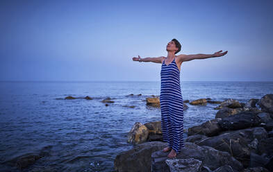 Happy mature woman standing barefoot on rocks in front of the sea looking up, Italy - DIKF00348