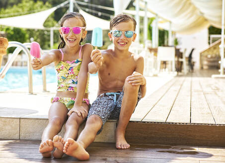 Porträt eines glücklichen kleinen Mädchens und eines Jungen mit verspiegelter Sonnenbrille, die ihr Eis am Stiel zeigen - DIKF00359