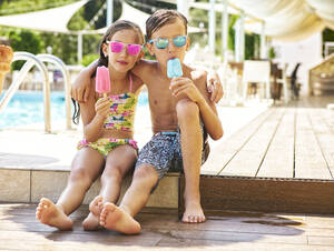 Porträt eines kleinen Mädchens und eines Jungen mit Eis am Stiel und verspiegelter Sonnenbrille vor einem Schwimmbad - DIKF00360
