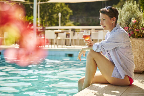 Reife Frau sitzt am Pool mit einem Glas Rotwein - DIKF00363