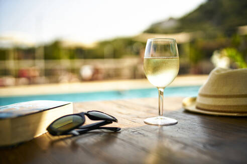 Glass of cooles white wine, book, sunglasses and straw hat in front of swimming pool, Italy - DIKF00366
