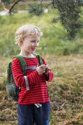 Porträt eines lächelnden blonden Jungen mit Rucksack in der Natur - IHF00235