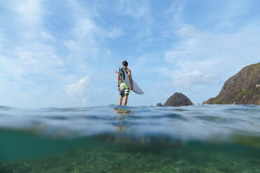 Surfer holding his surf board, standing on stone, Sumbawa island, Indonesia, over-under image - KNTF03858