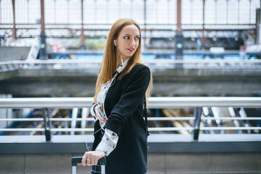 Junge Frau mit Kopfhörern und Mobiltelefon, die sich am Bahnhof umdreht - KIJF02870