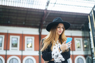 Junge Frau mit Hut benutzt ein Handy am Bahnhof - KIJF02883
