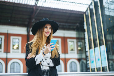 Lächelnde junge Frau mit Hut, die am Bahnhof ein Mobiltelefon benutzt - KIJF02884