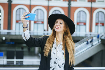 Smiling young woman wearing a hat taking a selfie at train station - KIJF02885