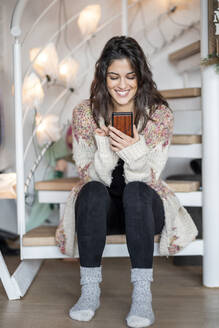 Porträt einer glücklichen jungen Frau, die zu Hause auf der Treppe sitzt und ihr Smartphone benutzt - FMKF06053
