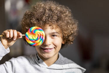 Portrait of smiling boy with brown ringlets covering eye with colourful lollipop - FMKF06057
