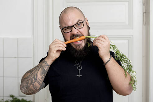 Portrait of bearded man biting in carrot in kitchen - FLLF00373