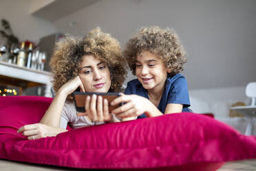 Mutter und Sohn sehen sich ein Video auf dem Smartphone an und liegen auf einem großen Kissen - FMKF06068