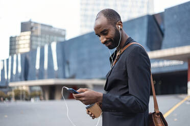 Portrait of smiling young businessman with coffee to go listening music with earphones and smartphone - JSRF00693