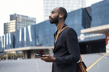 Young businessman with listening music with earphones and smartphone outdoors - JSRF00694