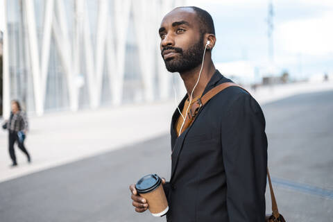 Porträt eines jungen Geschäftsmannes mit Kaffee zum Mitnehmen, der mit Kopfhörern im Freien Musik hört, lizenzfreies Stockfoto