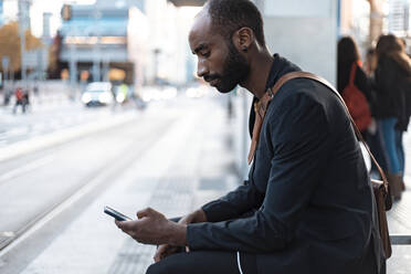 Junger Geschäftsmann sitzt an der Straßenbahnhaltestelle und schaut auf sein Smartphone - JSRF00702