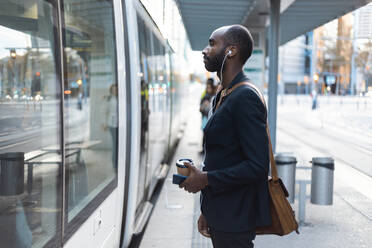 Junger Geschäftsmann mit Kaffee zum Mitnehmen steht an der Straßenbahnhaltestelle und hört Musik mit Kopfhörern und Smartphone - JSRF00704