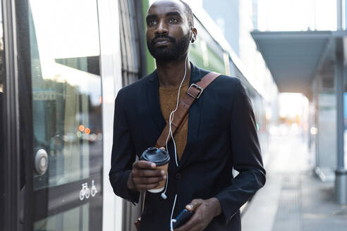 Porträt eines jungen Geschäftsmannes mit Coffee to go, Kopfhörern und Smartphone an einer Straßenbahnhaltestelle - JSRF00705