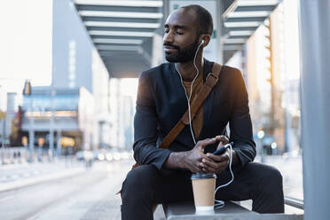 Junger Geschäftsmann mit Kaffee zum Mitnehmen, der mit Kopfhörern und Smartphone Musik hört und sich an einer Straßenbahnhaltestelle entspannt - JSRF00706