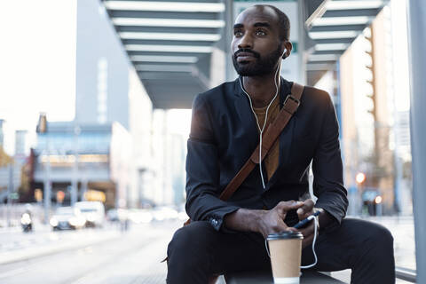 Porträt eines jungen Geschäftsmannes, der mit Kopfhörern und Smartphone an einer Straßenbahnhaltestelle Musik hört, lizenzfreies Stockfoto