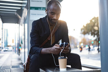Junger Geschäftsmann sitzt abends an der Straßenbahnhaltestelle und benutzt Kopfhörer und Smartphone - JSRF00708