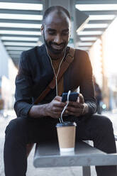 Smiling young businessman sitting at tram stop using earphones and smartphone - JSRF00709