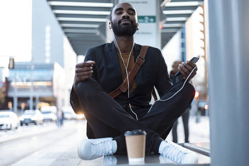 Portrait of young businessman with earphones and smartphone meditating on bench at tram stop - JSRF00710