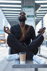 Portrait of young businessman with earphones and smartphone meditating on bench at tram stop - JSRF00711