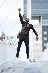 Happy young businessman with coffee to go, smartphone and earphones jumping in the air - JSRF00715