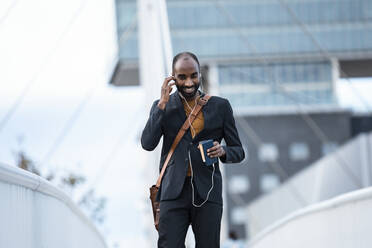 Lächelnder junger Geschäftsmann mit Smartphone und Kopfhörern im Freien - JSRF00716