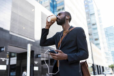 Young businessman with earphones and smartphone drinking coffee to go outdoors - JSRF00721