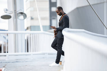Junger Geschäftsmann mit Kopfhörern steht auf einem Steg und schaut auf sein Smartphone - JSRF00723