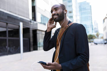 Smiling young businessman listening music with earphones and smartphone in the city - JSRF00727