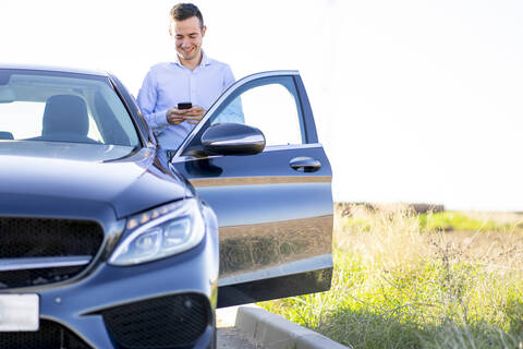 Lächelnder junger Geschäftsmann, der sein Smartphone am Auto auf der Landstraße benutzt, lizenzfreies Stockfoto