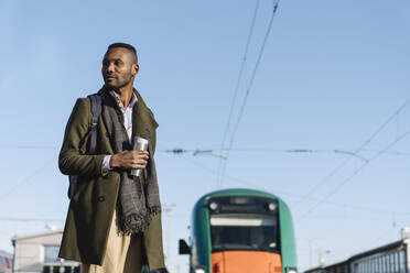 Porträt eines stilvollen Mannes mit wiederverwendbarem Becher, der auf den Zug wartet - AHSF01630