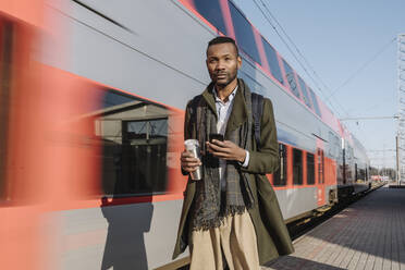 Porträt eines stilvollen Mannes mit Smartphone neben einem Zug - AHSF01634