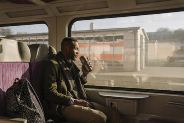 Stilvoller Mann trinkt während einer Zugfahrt ein Heißgetränk aus einem wiederverwendbaren Becher - AHSF01638