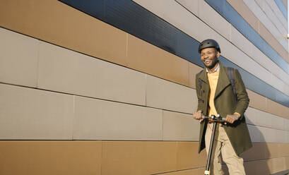 Happy man with helmet and scooter passing a building - AHSF01659