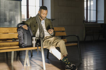 Stylish man using smartphone while sitting on a bench in a train station - AHSF01677