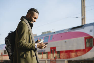 Stilvoller Mann mit wiederverwendbarem Becher und Smartphone beim Warten auf den Zug - AHSF01679