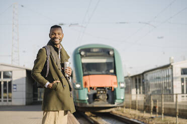 Porträt eines lächelnden Mannes mit wiederverwendbarem Becher, der auf den Zug wartet - AHSF01685