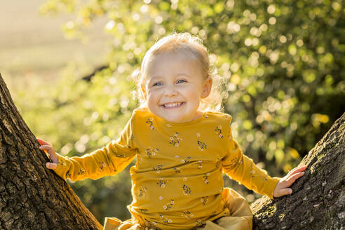 Portrait of happy blond toddler girl sitting on tree trunk - NMSF00381