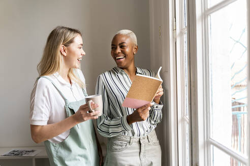 Zwei glückliche Frauen mit Notizbuch und Tasse Kaffee am Fenster - AFVF04757