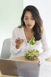 Portrait of young architect eating mixed salad at desk looking at laptop - AFVF04772